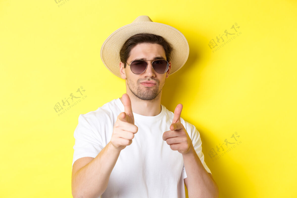 时尚旅游和度假的概念一个戴着夏天帽子和太阳镜的帅哥的特写镜头 用手指指着镜头 站在黄色背景上帽子自信太阳镜