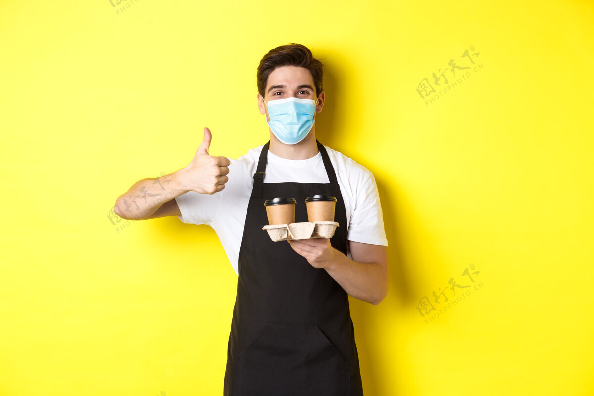 咖啡师covid-19的概念 咖啡馆和社交距离戴着医用面具和黑色围裙的年轻男咖啡师拿着外卖咖啡杯 竖起大拇指 黄色背景男士外科面膜表情