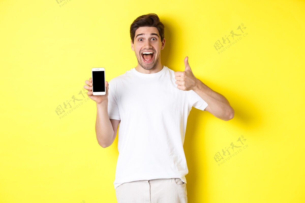 竖起大拇指快乐的年轻人展示拇指和手机屏幕 推荐应用程序或互联网站立男人帅气