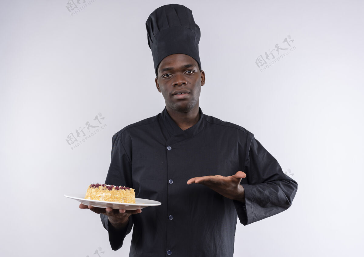 持有身着厨师制服的年轻自信的美国黑人厨师拿着并指着白色盘子上的蛋糕 上面留有复印空间非裔美国人信心蛋糕