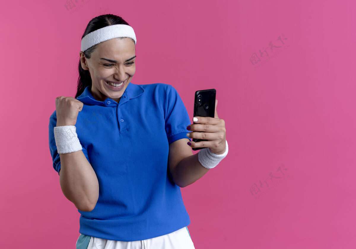 年轻年轻快乐的白人运动女性戴着头带和腕带看着手机 举起拳头放在粉色和复印空间上粉红色空间拳头