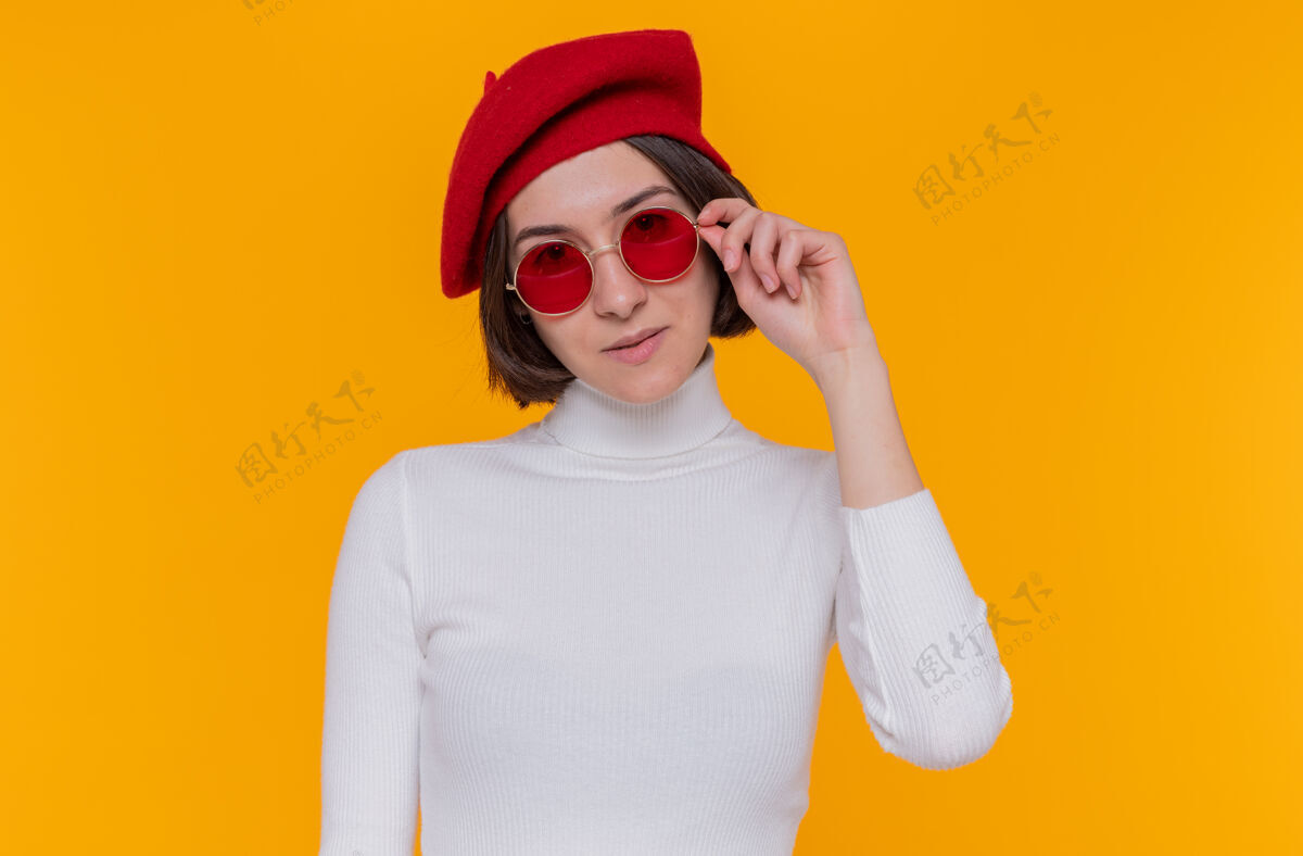 头发身穿白色高领毛衣 头戴贝雷帽 戴着红色太阳镜 留着短发的年轻女子站在橙色的墙上 面带微笑 自信 快乐 积极地看着前方年轻微笑贝雷帽
