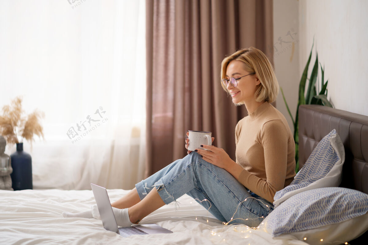 阅读女人在卧室里用笔记本电脑放松 喝一杯热咖啡或茶电脑交流床