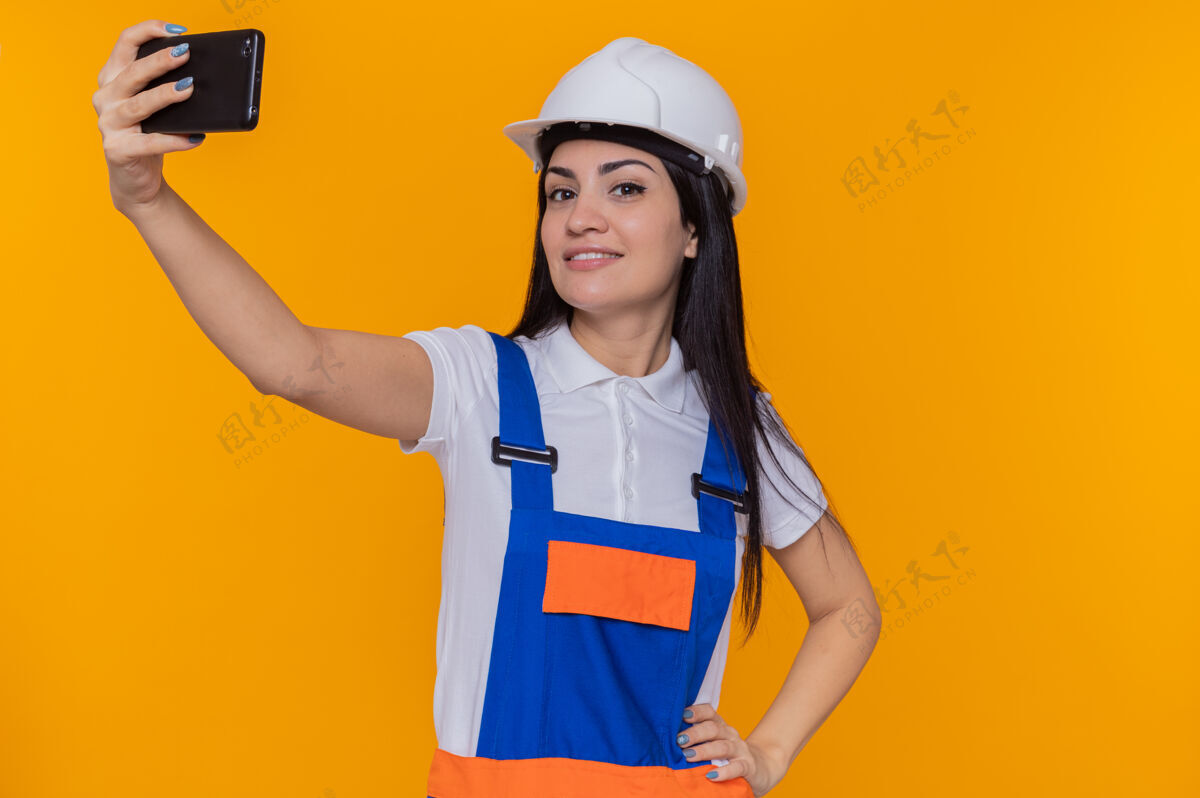 制服身穿施工制服 头戴安全帽的年轻建筑工人正在用智能手机自拍 自信地微笑着站在橙色的墙上头盔站立微笑