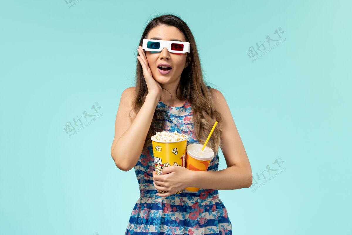 年轻女性正面图年轻的女性拿着爆米花 戴着d墨镜在蓝色的表面上喝酒爆米花漂亮性感