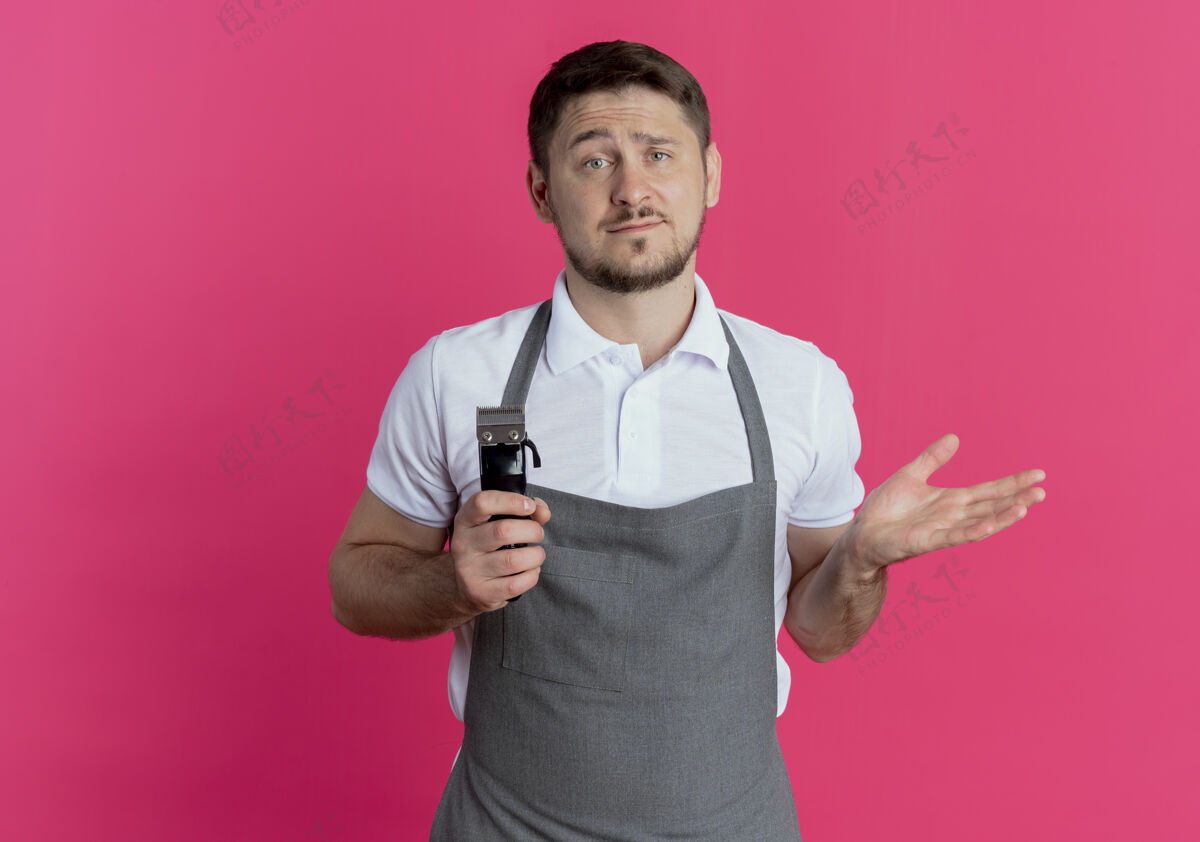 男人围裙上的理发师手里拿着剃须刀站在粉红色的墙上 困惑而不快修剪困惑不高兴
