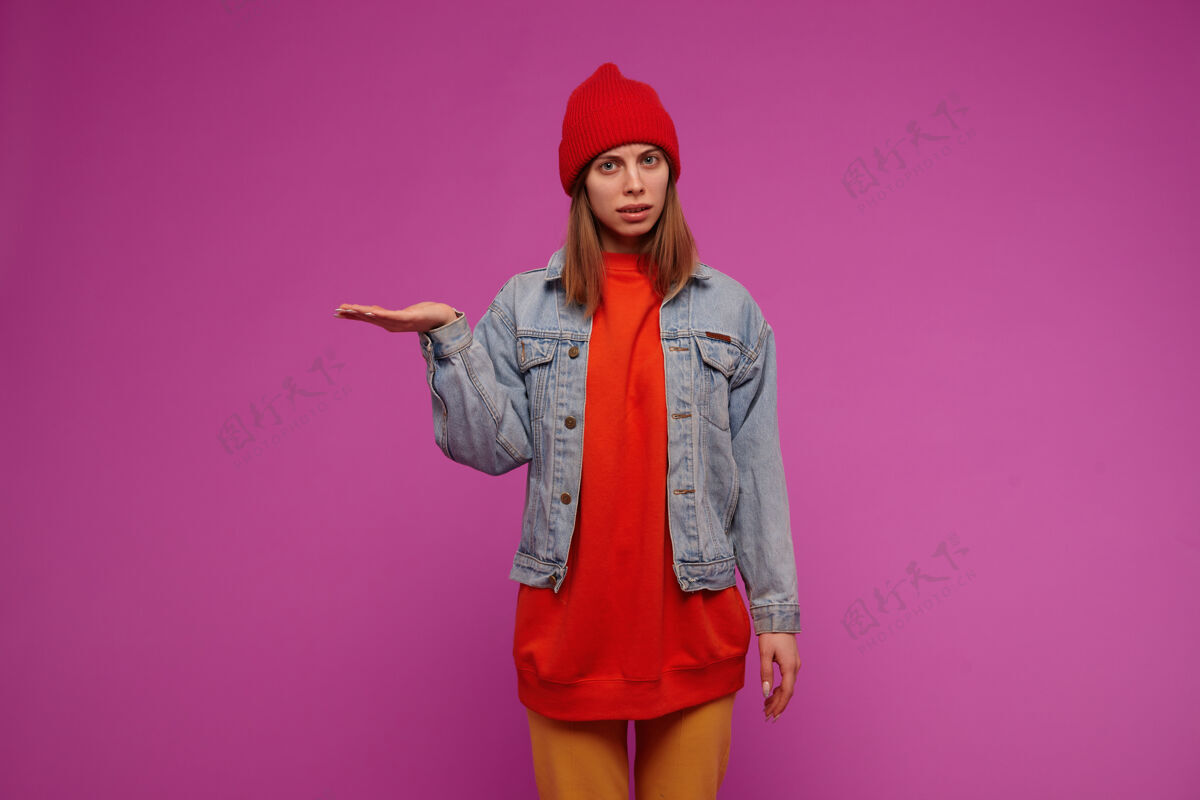 年轻人漂亮漂亮的女孩 深色头发穿着牛仔裤夹克 黄色裤子 红色毛衣和帽子假装拿着什么东西在紫色的墙上学生年轻人20多岁