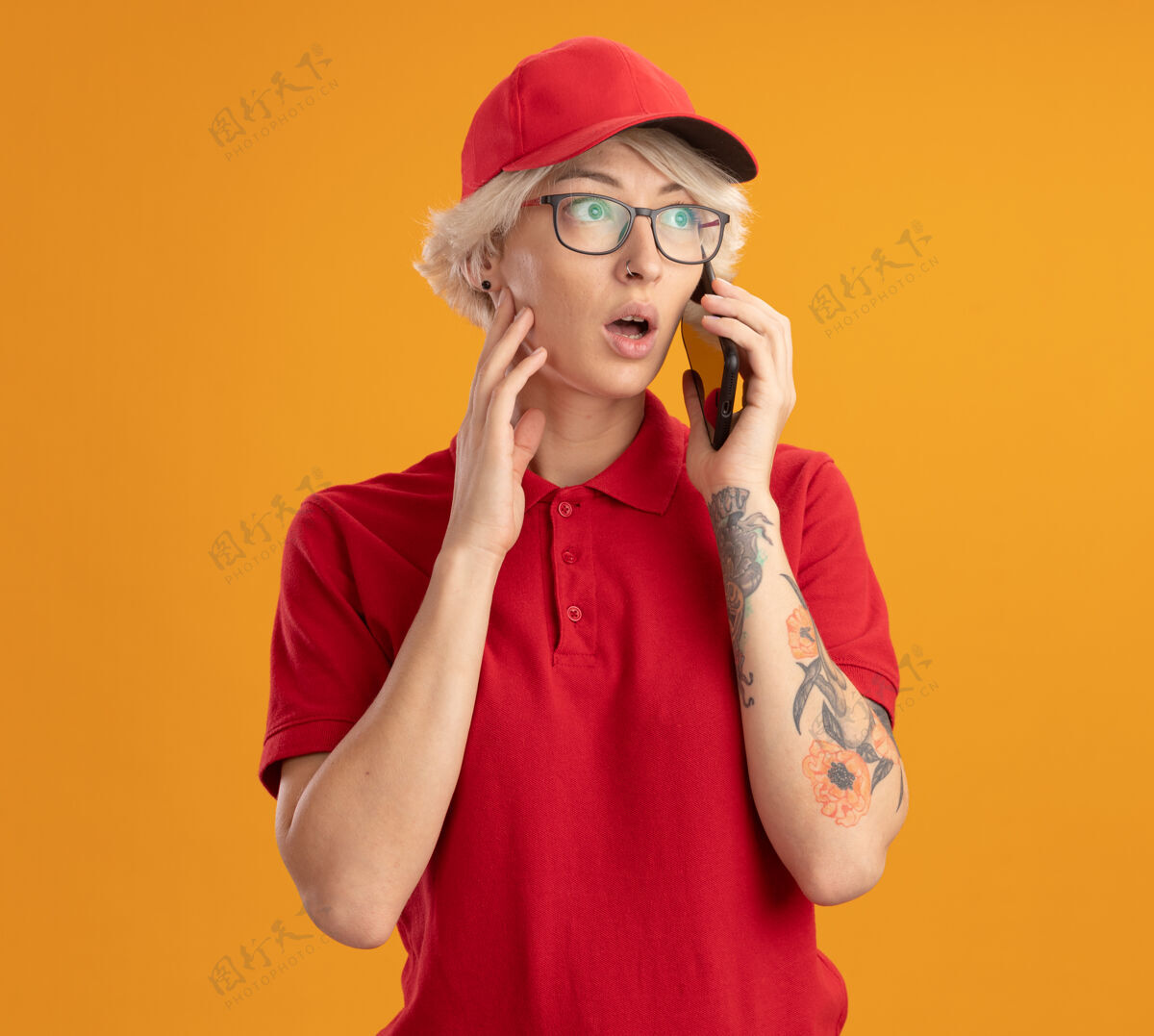 非常身穿红色制服 戴着眼镜的年轻女送货员站在橙色的墙上讲着手机 感到困惑和焦虑电话女人制服