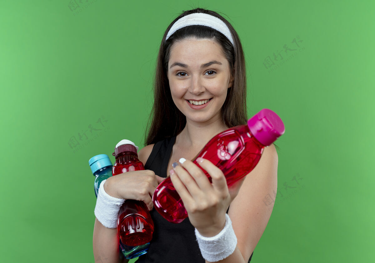 头带戴着头巾的年轻健身女 手里拿着几瓶水 站在绿色的墙边微笑着给其中一个送水提供微笑抱着