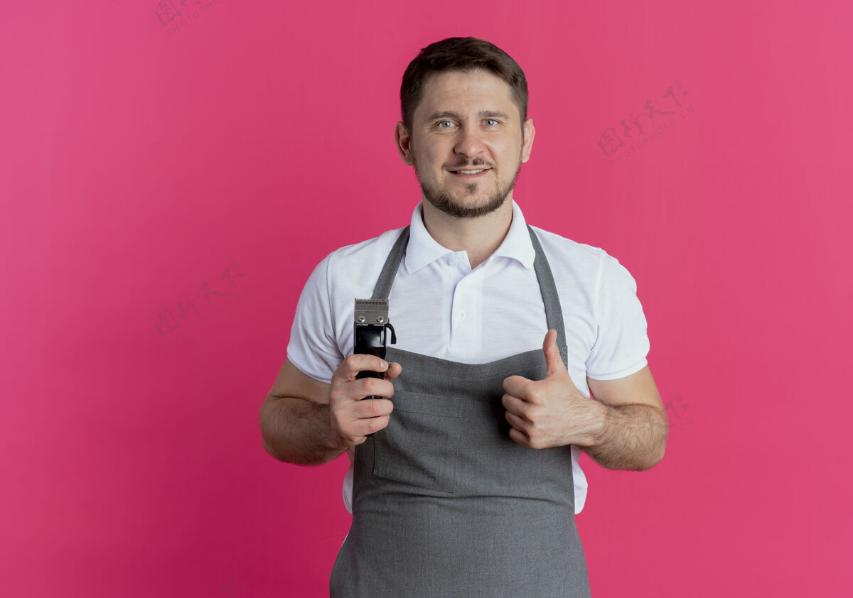 站着围裙上的理发师拿着剃须刀 竖起大拇指微笑着站在粉红色的墙上拿着围裙修剪
