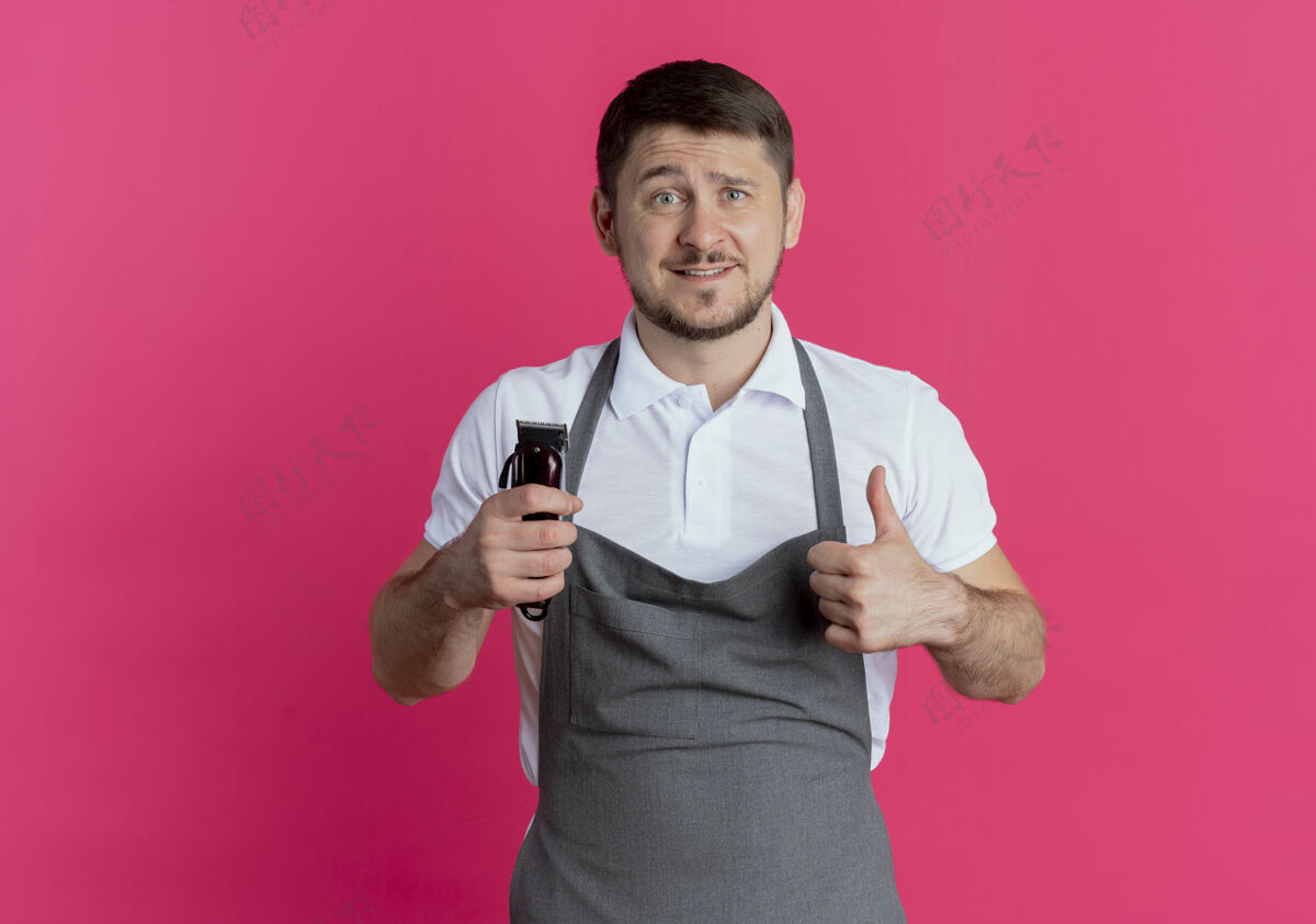 男人围裙上的理发师拿着剃须刀 竖起大拇指微笑着站在粉红色的墙上大拇指站着拿着