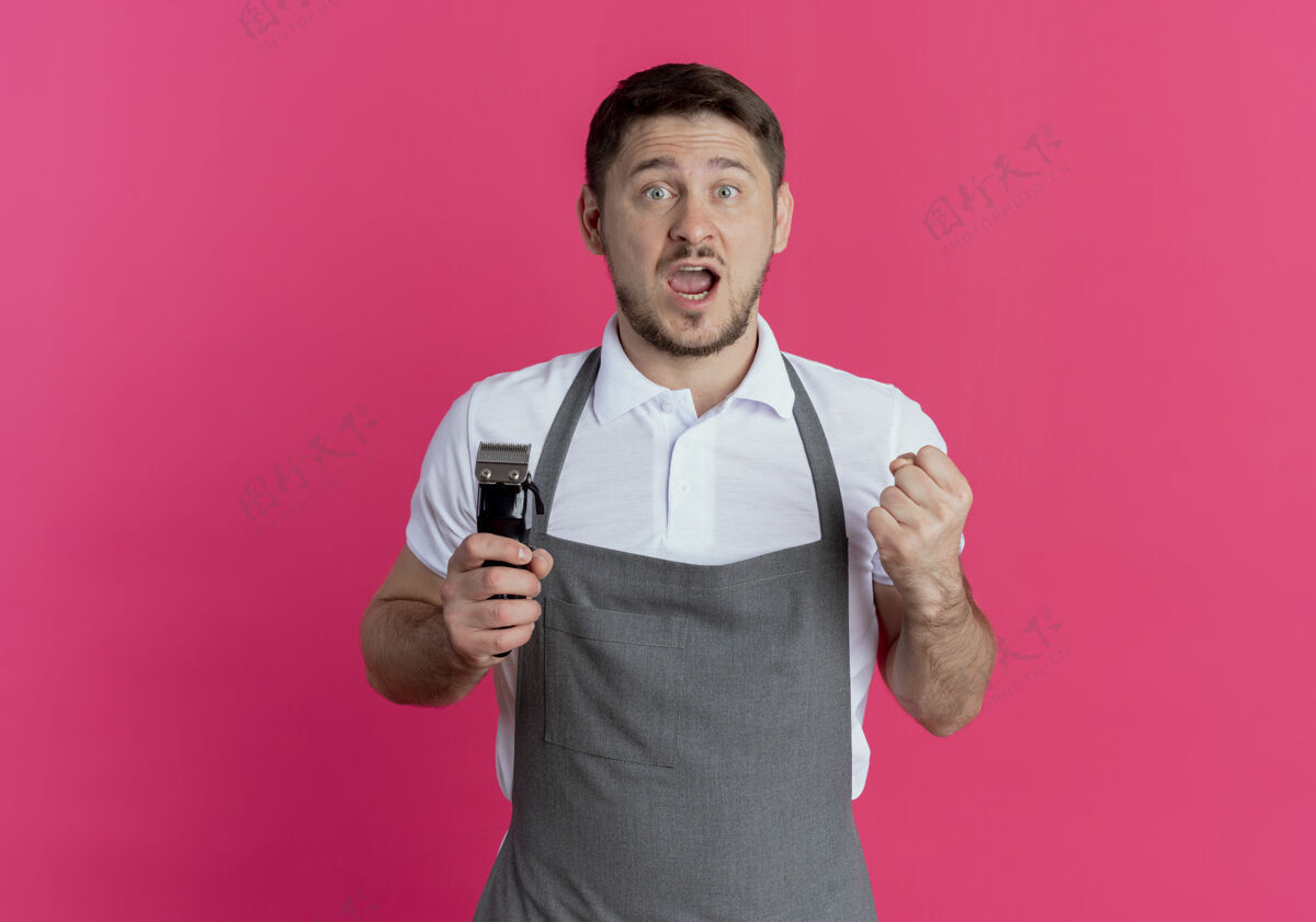 理发师围裙上的理发师手里拿着剃须刀紧握着拳头站在粉红色的墙上 高兴又兴奋男人站着握紧