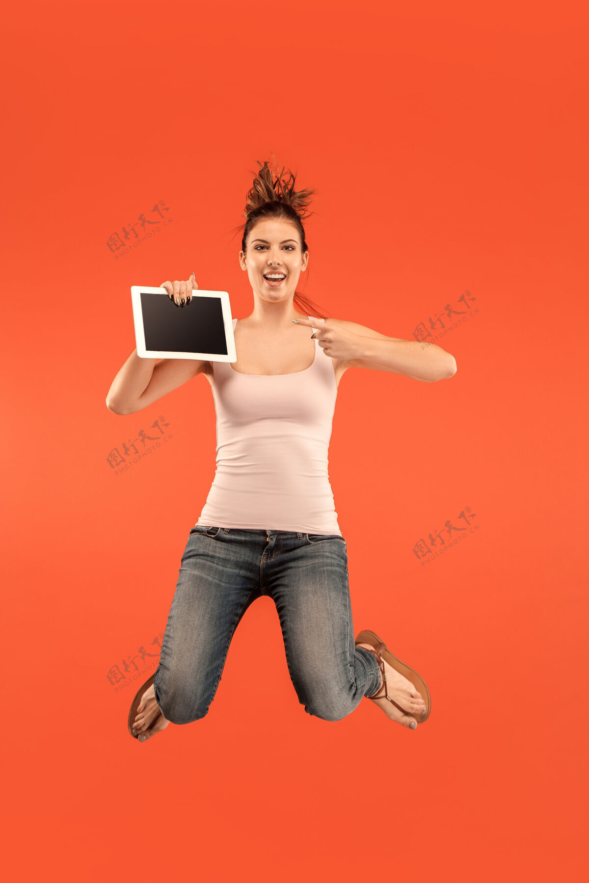 平板电脑年轻女子在蓝色工作室背景上跳跃时使用笔记本电脑或平板电脑小玩意在运动或运动中奔跑的女孩休闲运动前面