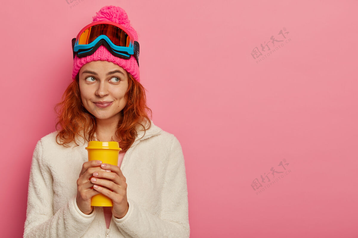 咖啡迷人的红发女人戴着暖和的帽子 滑雪镜和白色套头衫一次性衣服满意