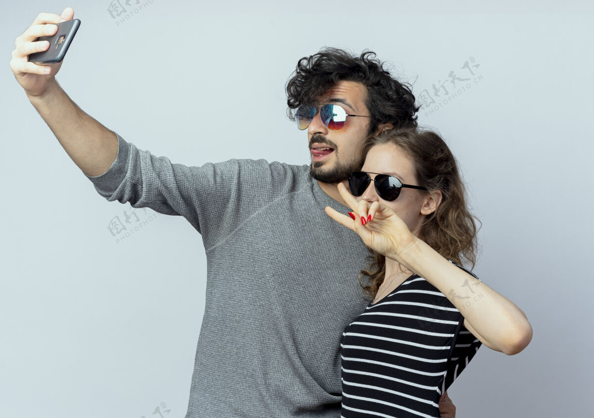 站着一对年轻的情侣热恋中 幸福的男人用智能手机拍下了他们站在白墙上的照片情侣拿着男人