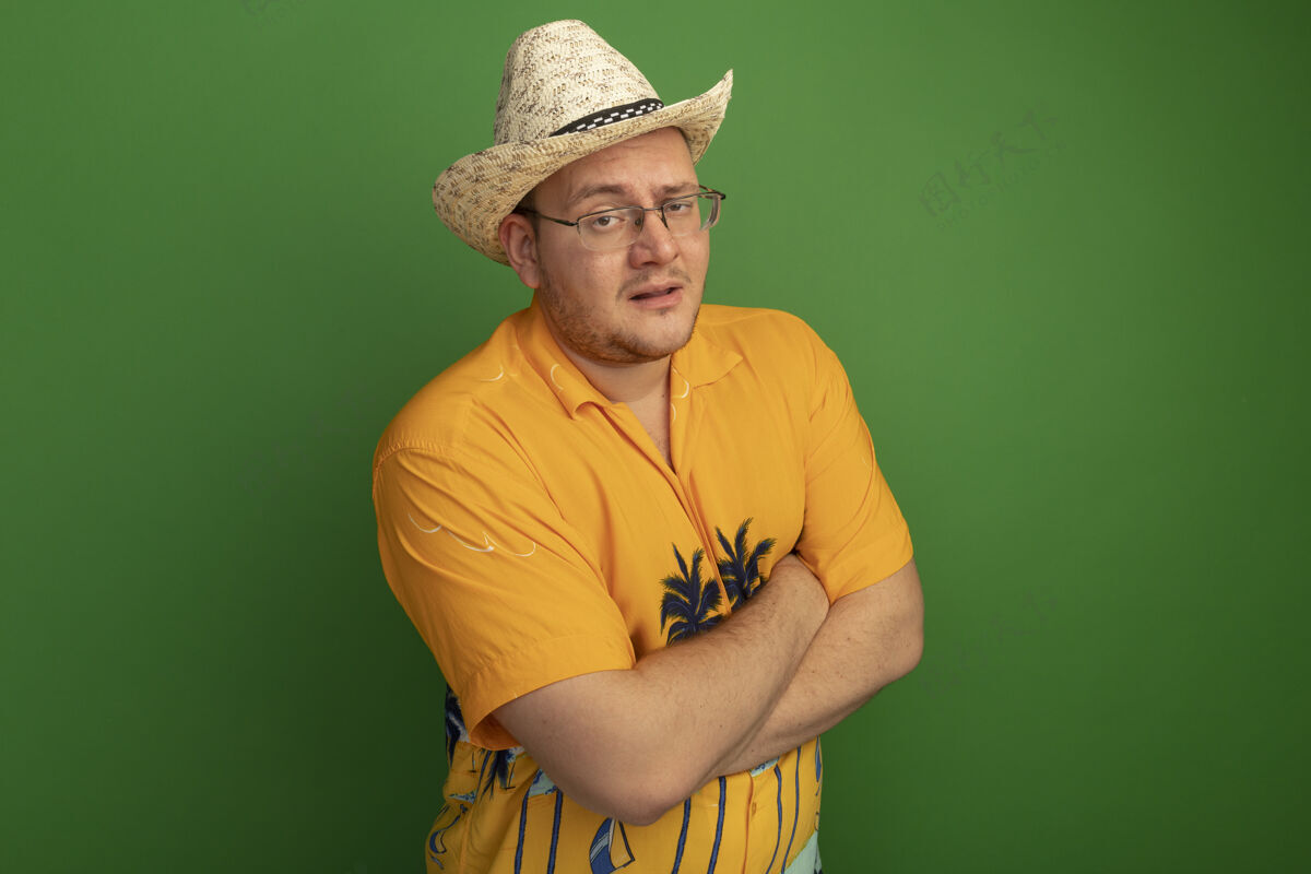 帽子戴着眼镜的男人穿着橙色衬衫 戴着夏令帽 严肃的脸交叉着双臂站在绿色的墙上穿夏天男人