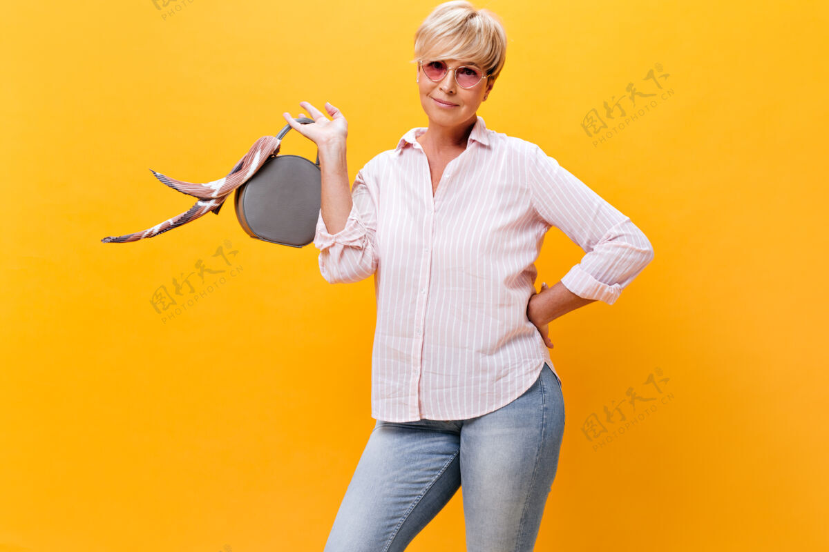 成熟穿着时髦服装戴墨镜的成年女士拿着手提包摆姿势橙色背景女士老年