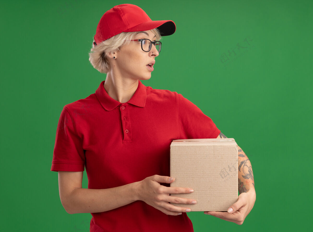 女人年轻的送货员身穿红色制服 戴着帽子 戴着眼镜 拿着纸板箱 站在绿色的墙上 表情困惑地看着一边站着抱着表情