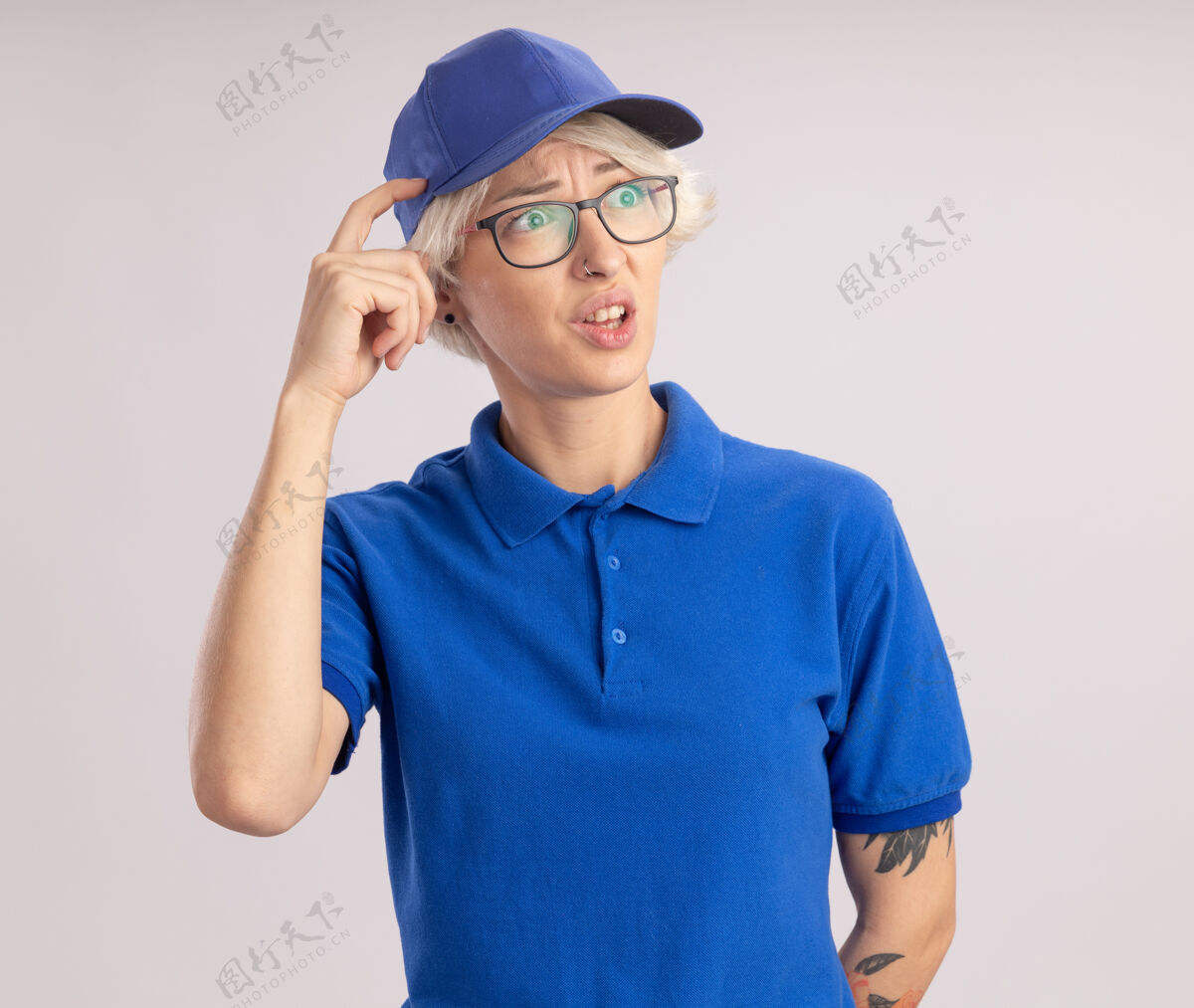 头穿着蓝色制服 戴着帽子的年轻女送货员一边看一边困惑地抓着头 手指站在白色的墙上制服抓伤帽子