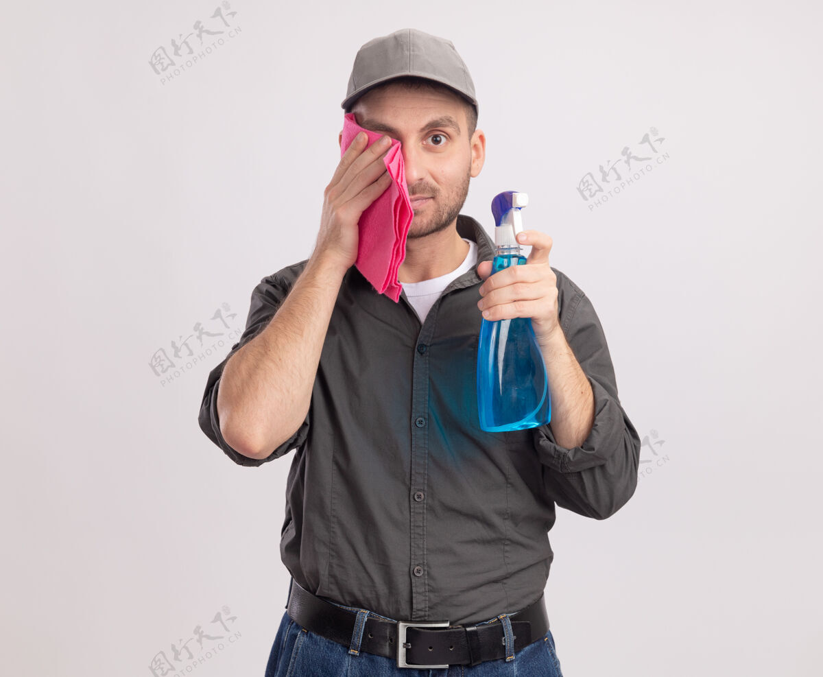 清洁年轻的清洁工穿着休闲服 戴着帽子 拿着清洁喷雾和抹布站在白色的墙上用抹布擦眼睛男人衣服磨损