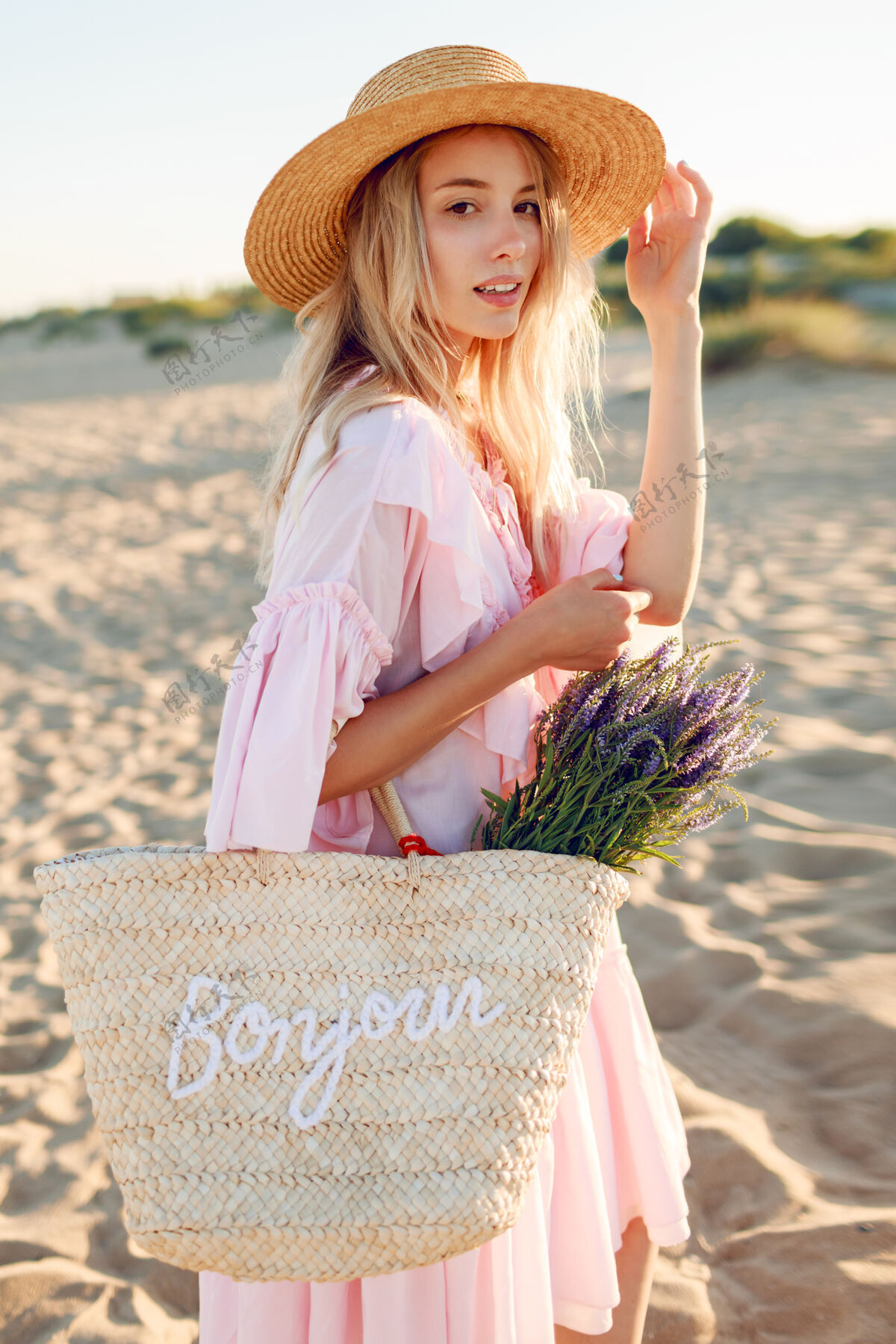 风浪漫的白人女子 戴着时髦的帽子 穿着优雅的粉色连衣裙 手持草包和花束 在沙滩上摆着姿势海滩薰衣草肖像
