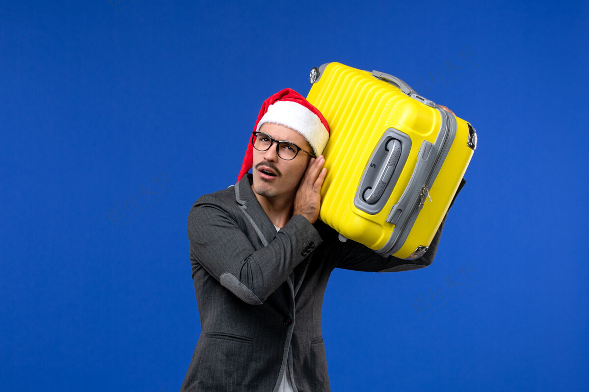 专业正面图年轻男子背着沉重的黄包在蓝墙飞机上度假包年轻男性头盔