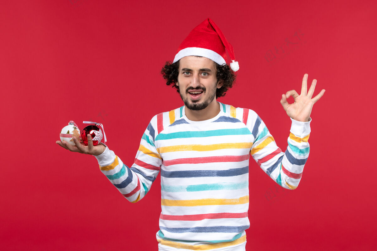 男性正面图年轻人手持圣诞树玩具在红墙新年红人节前面人帽子