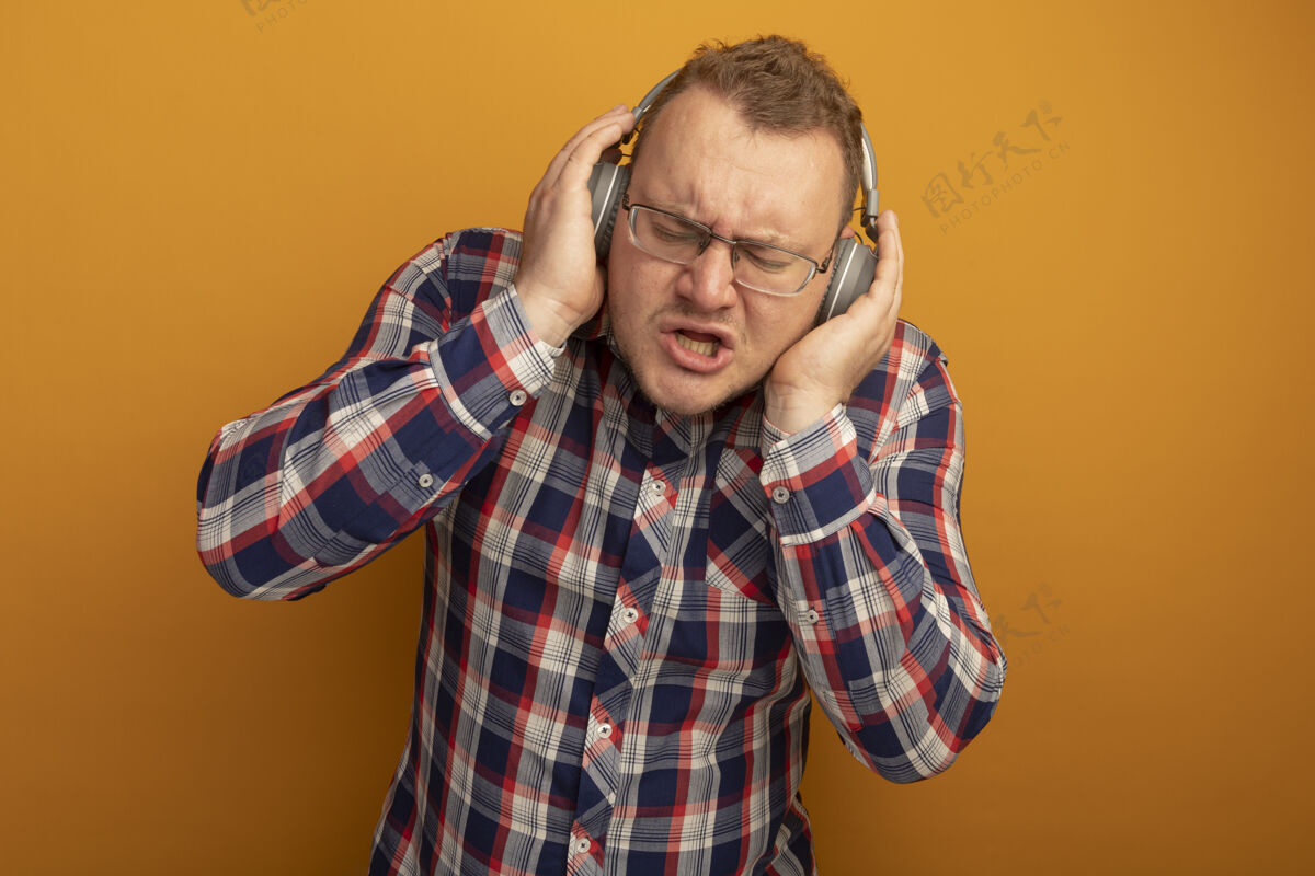 音乐戴着眼镜 穿着格子衬衫 戴着耳机的情绪化男人站在橙色的墙上欣赏音乐男人站立眼镜