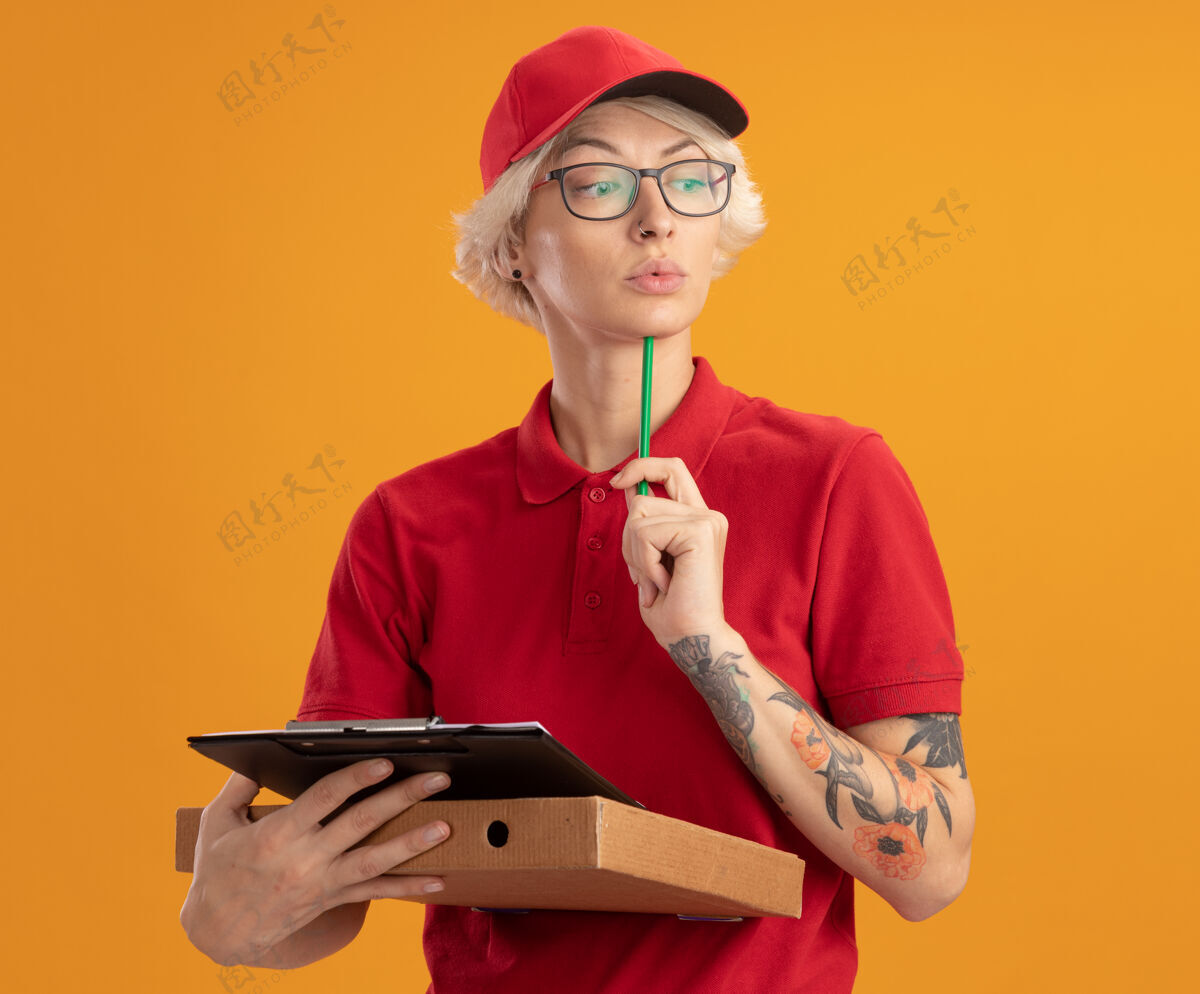 帽子身穿红色制服 戴着帽子 戴着眼镜 拿着披萨盒和铅笔的剪贴板的年轻女送货员站在橙色的墙上 神情严肃地站在一边思考剪贴板披萨拿着