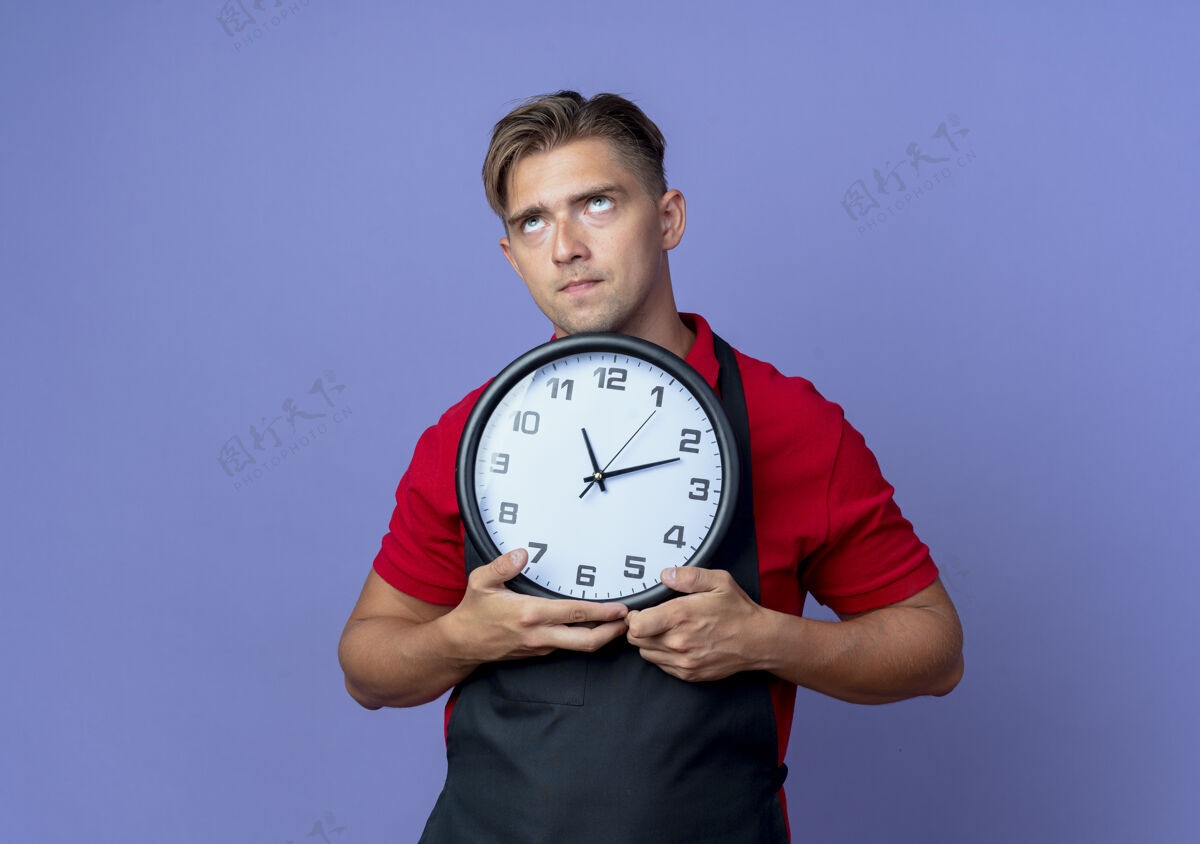 空间年轻体贴的金发男理发师穿着制服 拿着时钟 在紫色的空间和复印空间上孤立地看着男性向上看
