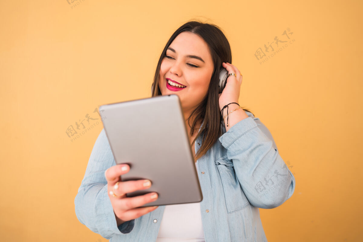 享受在黄色背景下 戴着耳机和数字平板电脑在户外听音乐的大号年轻女子的画像媒体耳机大小