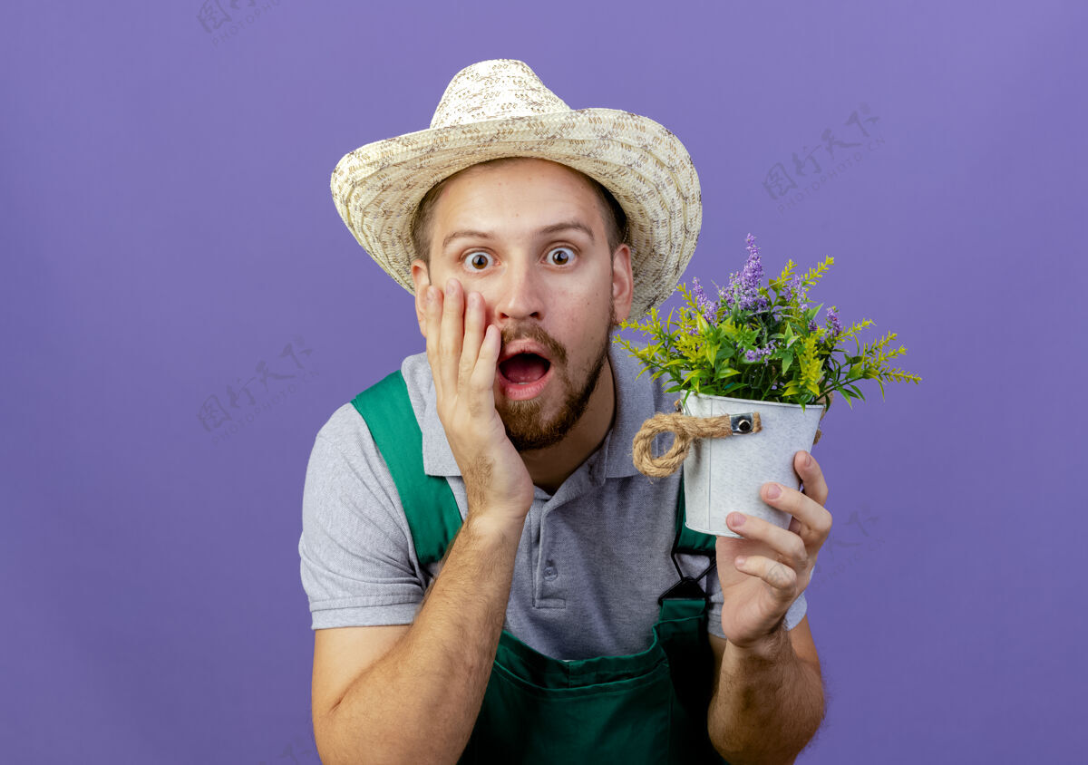 看着震惊年轻英俊的斯拉夫园丁制服和帽子举行花盆触摸脸孤立在紫色墙上复制空间斯拉夫脸花园