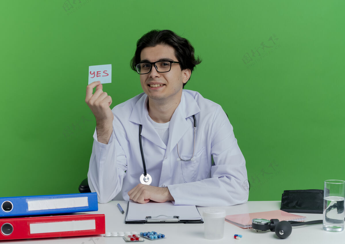 听诊器年轻的男医生穿着医用长袍 戴着听诊器 戴着眼镜 坐在办公桌旁 手里拿着医疗工具 绿墙上写着“是的”字样眼镜看男性