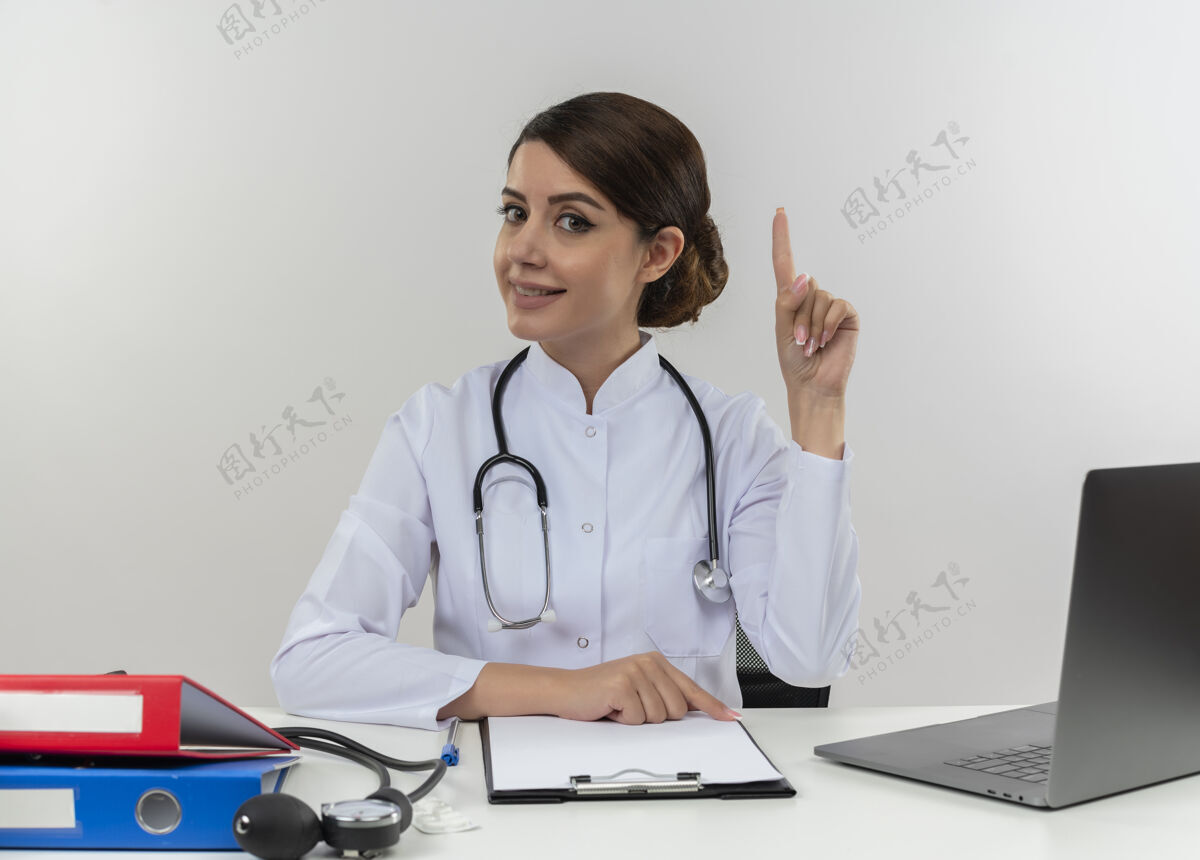 穿面带微笑的年轻女医生穿着医用长袍 听诊器坐在桌子旁 医疗工具和笔记本电脑孤立地指向白色的墙上医疗工具医生