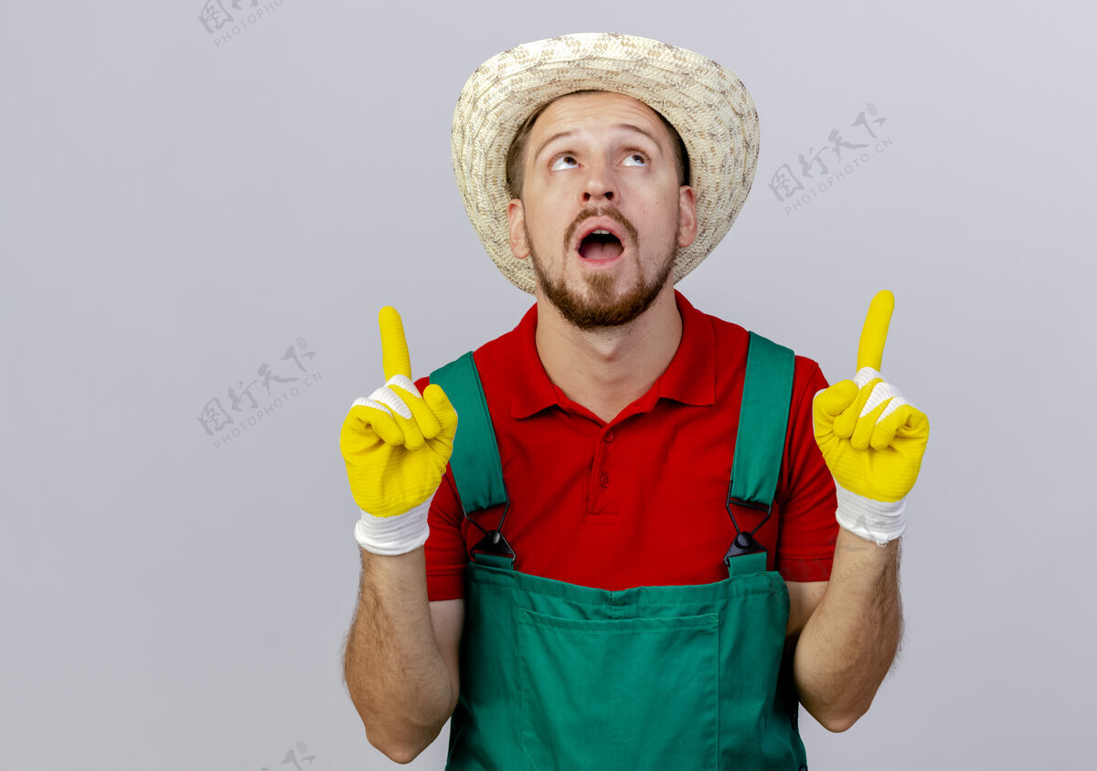 指向穿着制服的年轻英俊的斯拉夫园丁戴着园艺手套和帽子 看着并指着隔离在白色墙上的复制空间年轻花园手套