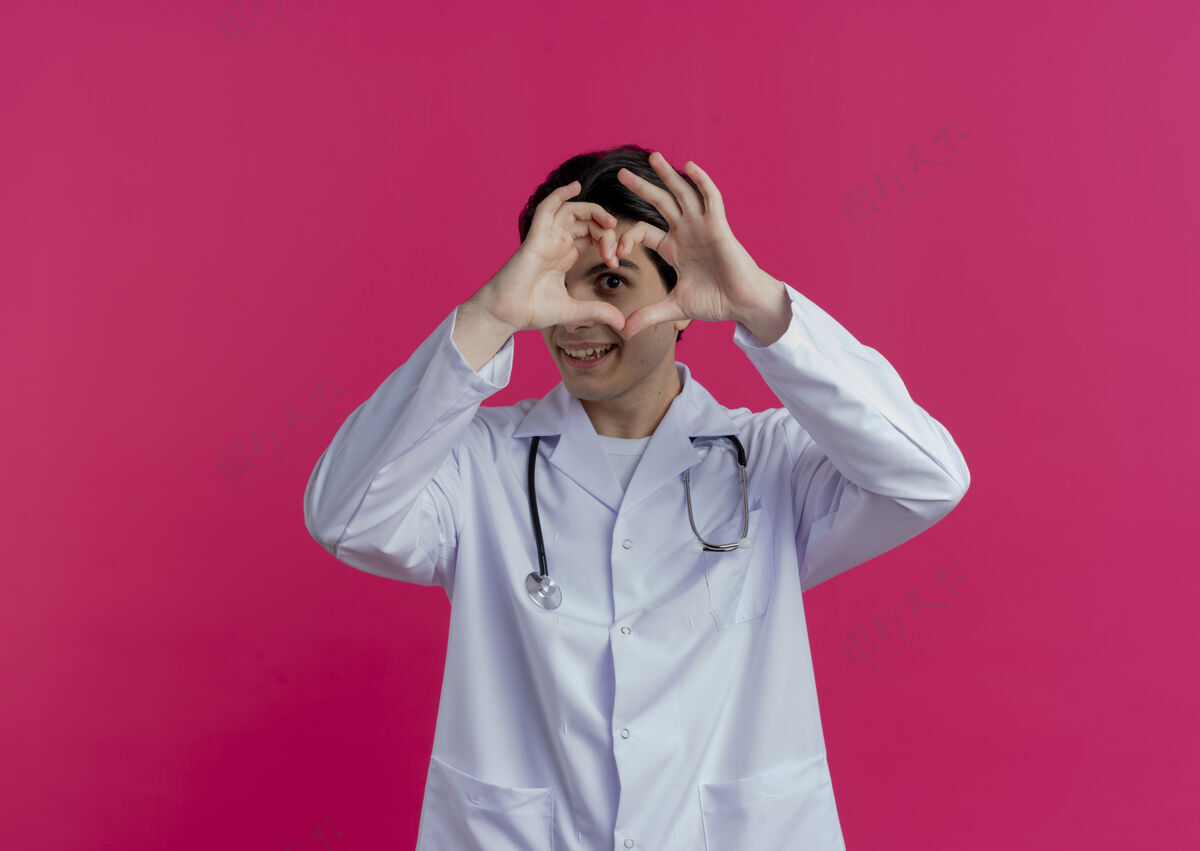 听诊器微笑的年轻男医生穿着医用长袍和听诊器在粉红色的墙上做心脏标志 并留有复印空间男性穿医生