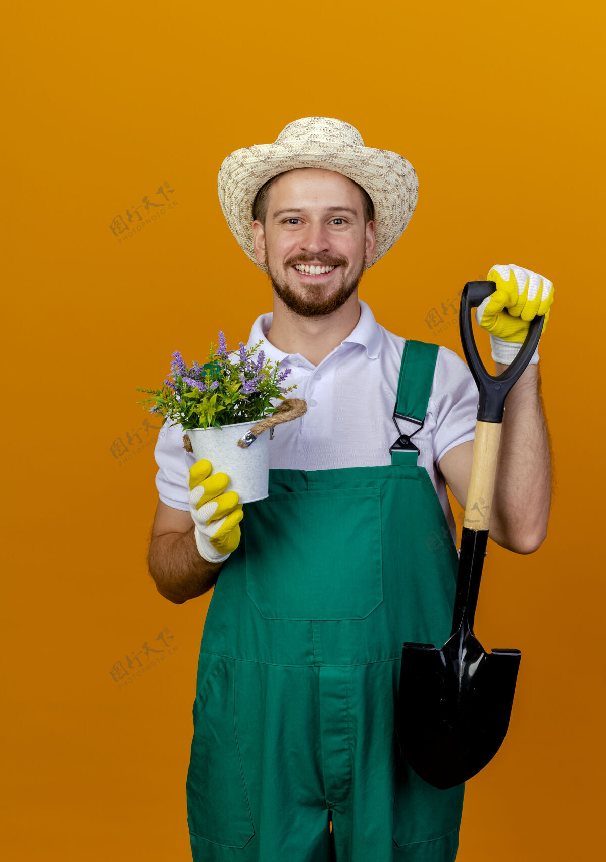 花园身着制服的年轻英俊的斯拉夫园丁面带微笑 戴着帽子和园艺手套 手里拿着铲子和花盆 看上去孤立在橙色的墙上橙色黑桃年轻人