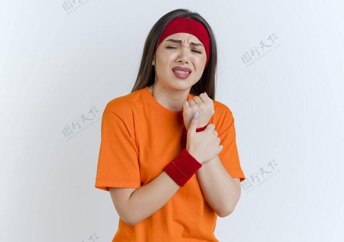 腕带疼痛的年轻运动女性戴着头带和腕带抱着手腕闭着眼睛隔离在白色的墙壁与复制空间运动抱着手腕