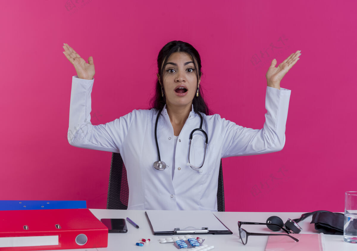 长袍令人印象深刻的年轻女医生穿着医用长袍和听诊器坐在办公桌上与医疗工具显示空手隔离在粉红色的墙上医疗听诊器年轻