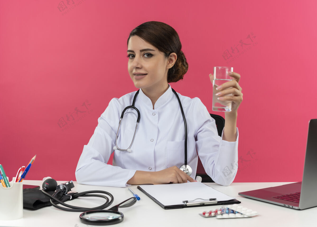 医生年轻的女医生穿着医用长袍 手持听诊器 坐在办公桌旁 手里拿着医疗工具和手提电脑 粉红色的墙上隔着一杯水女桌子长袍