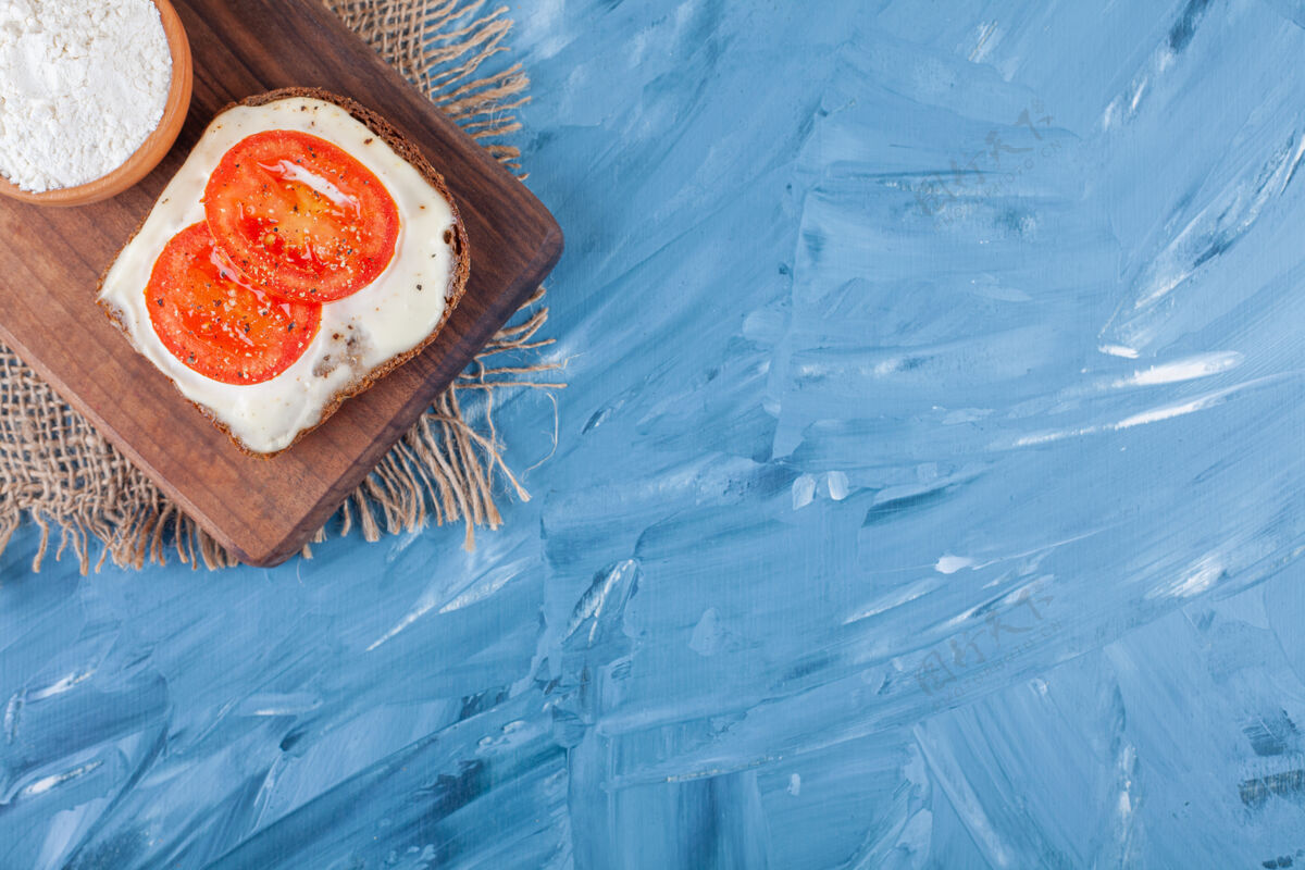 奶酪西红柿片放在奶酪面包上 一碗面粉放在切菜板上 粗麻布餐巾纸放在蓝色布上美味盐番茄