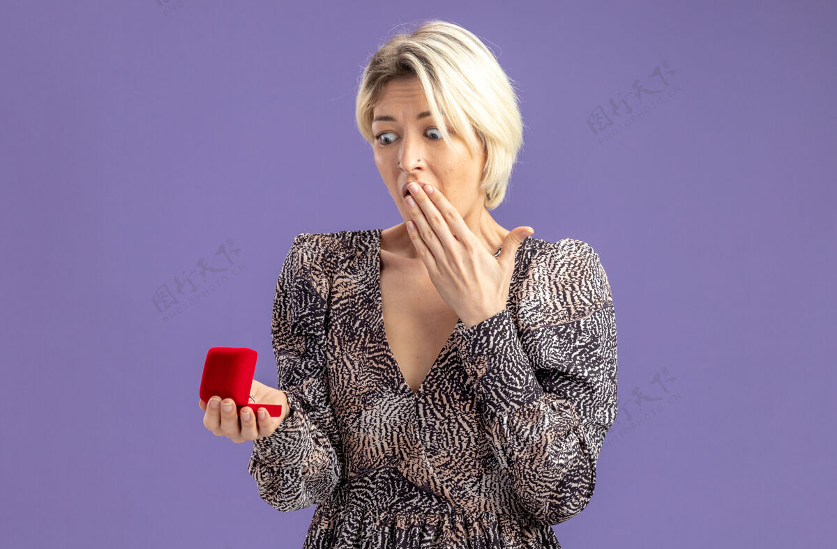 礼服年轻漂亮的女人穿着礼服拿着红色的订婚戒指看着它惊讶和惊喜的情人节概念站在紫色的墙上情人节立场女人