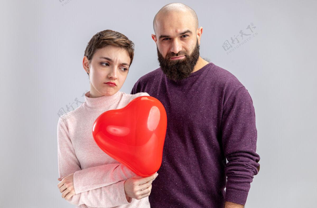 冒犯一对穿着休闲服的年轻夫妇冒犯了站在白墙上的留着胡须 留着短发 手持心形气球庆祝情人节的男女情人节立场衣服