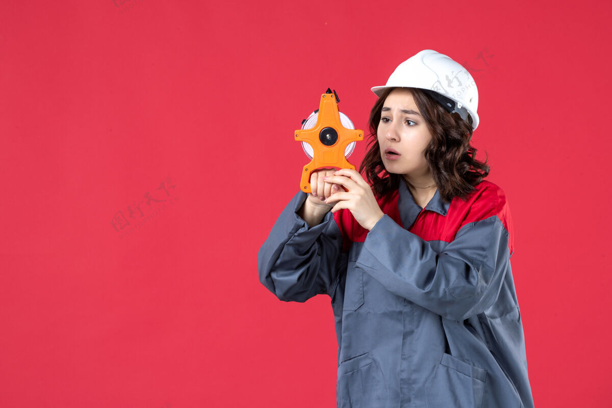 人前视图困惑的女建筑师穿着制服 戴着安全帽拿着卷尺 把注意力集中在隔离的红墙上微笑抱着测量