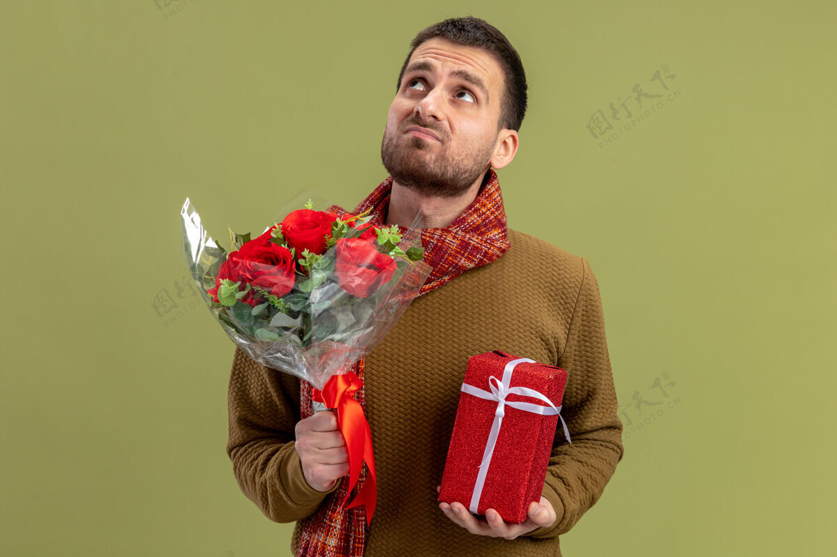 男人身穿休闲服的年轻人脖子上围着围巾 手里捧着一束红玫瑰 脸上带着悲伤的表情 站在绿色的墙上 端详着情人节的概念休闲围巾看着