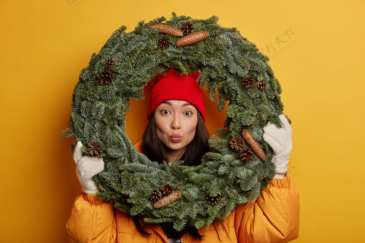 人可爱的韩国年轻女子嘴唇圆润 戴着绿色的云杉花环 穿着黄色的外套和白色的手套 在圣诞节前装饰房子 在室内摆姿势花圈站水平