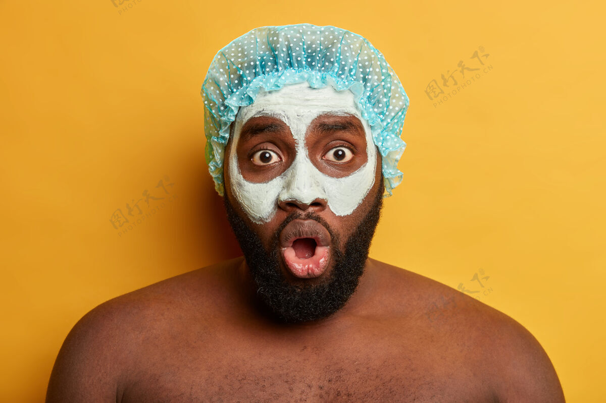 清洁震惊的黑皮肤没刮胡子的家伙脸上戴着粘土面具 浴帽 瞪大眼睛盯着镜头 有美容护肤的概念肖像卫生粘土