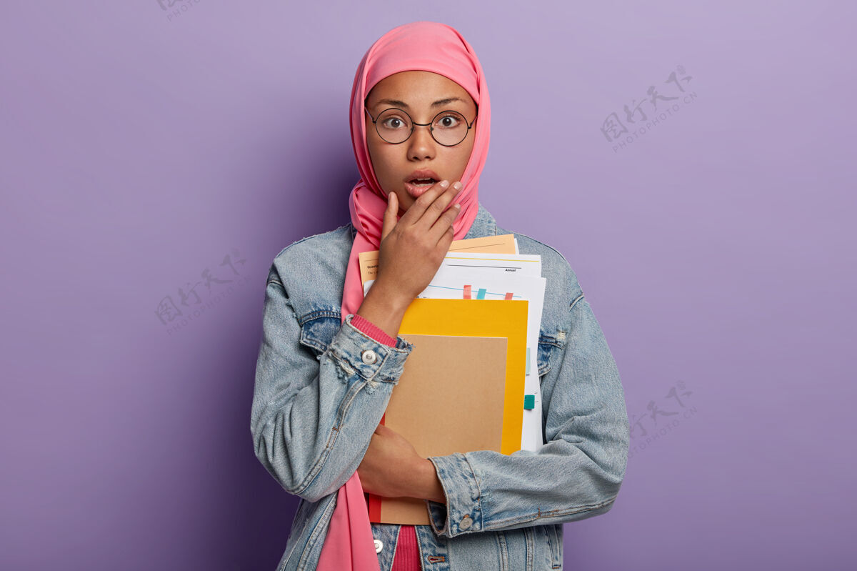 尴尬带着头巾的深肤色穆斯林妇女激动得目瞪口呆 惊讶地张开嘴 手里拿着一些纸和记事本昏迷信仰年轻