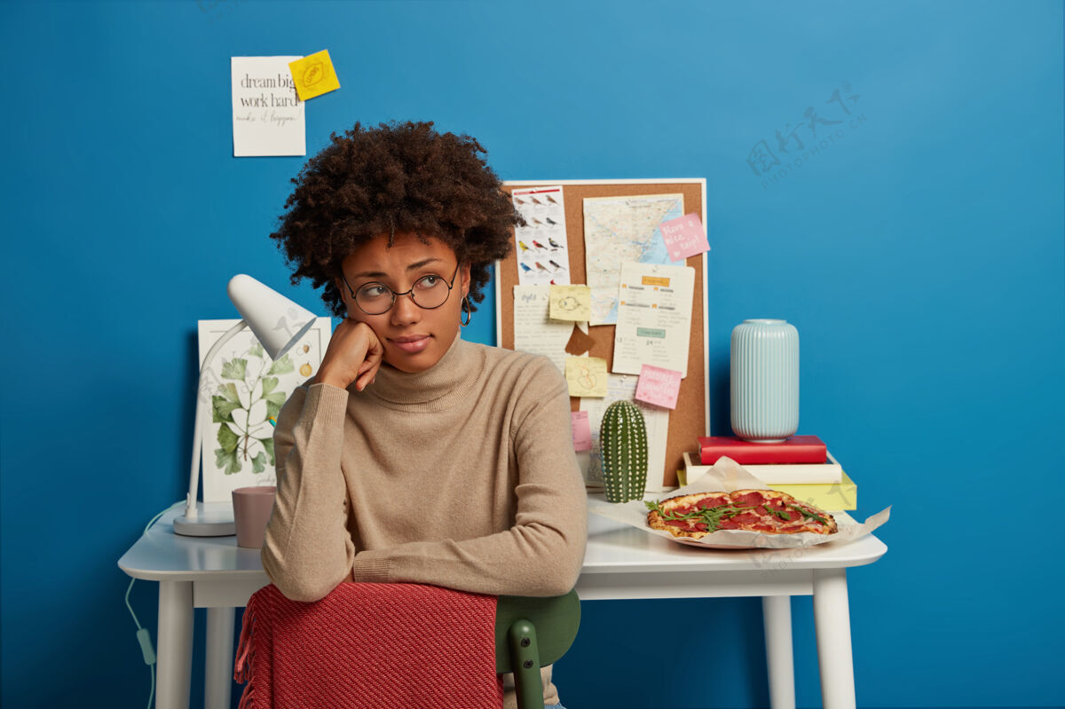 坐着悲伤的女人留着非洲式发型坐在椅子上 戴着圆眼镜和米黄色套头衫 坐在同事的空间里 桌子后面放着笔记本台灯和美味的比萨饼非洲房子工人