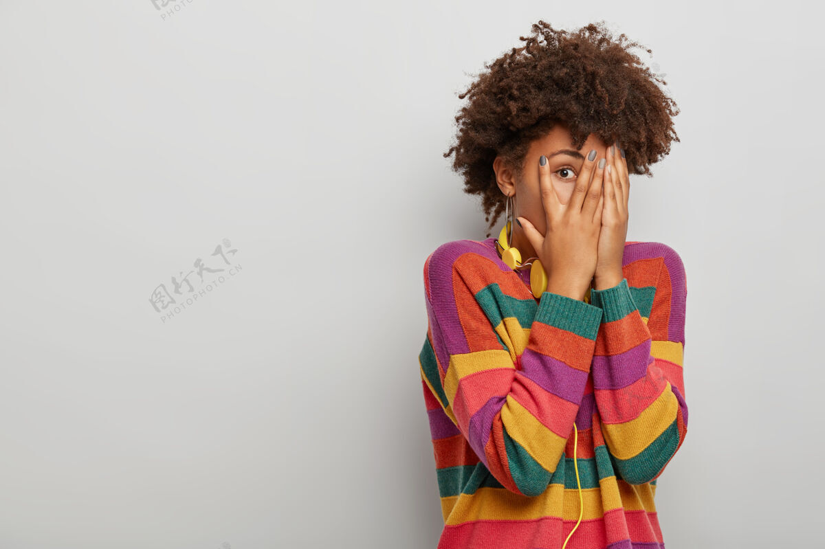 发型一个留着非洲发型的成年女人 用手指看 双手捂住脸 戴着耳机和条纹毛衣 站在白色的工作室背景下发型人偷看